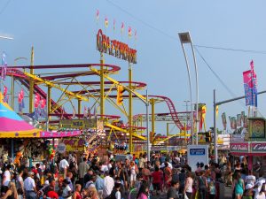 Daytime crowds packed along the Canadian National Exhibition midway in Toronto, with roller coaster track, game stalls and ride flags overhead.