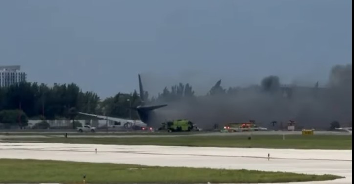 Aircraft rescue and firefighting trucks spraying the wreckage of a burning freighter across an airfield, the aircraft tail and a wing visible through heavy grey smoke.