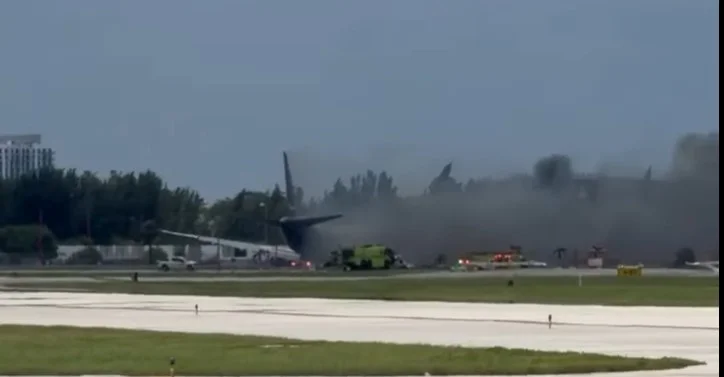 Aircraft rescue and firefighting trucks spraying the wreckage of a burning freighter across an airfield, the aircraft tail and a wing visible through heavy grey smoke.