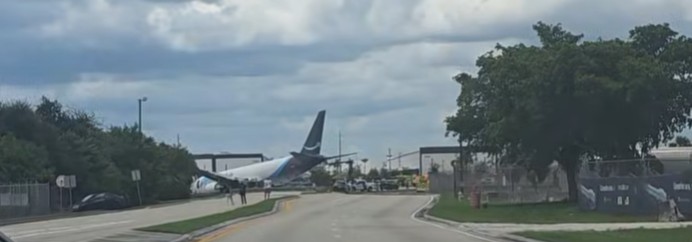 The tail fin of a white Boeing 767 freighter standing above a perimeter fence at the end of a public road, with stopped cars, people on the roadway and emergency vehicles alongside.