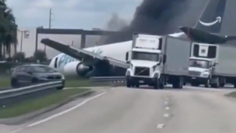A Boeing 767 freighter in Amazon Prime Air livery resting across a road at the edge of Miami International Airport, one wing extended over the roadway beside two struck transport trucks and a car, with black smoke rising behind the tail.