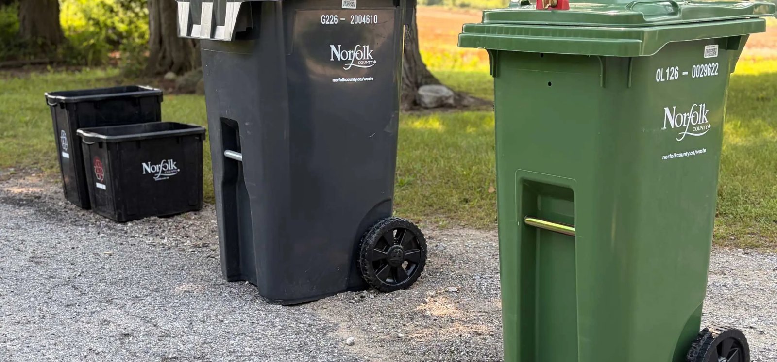 A grey Norfolk County garbage cart and a green organics cart at the end of a gravel driveway, with two black recycling boxes behind them