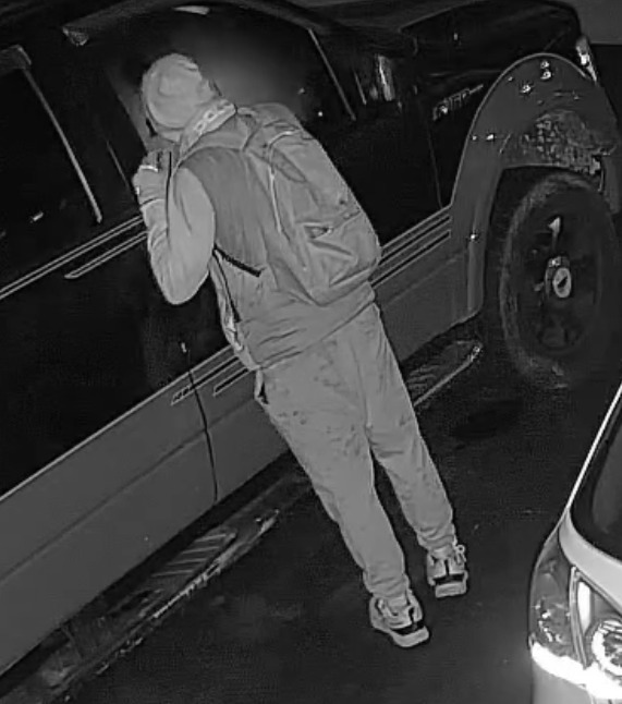 Peel police suspect image 2 — 32 vehicle break-ins, Mississauga (NR 26-170) Security-camera still released by Peel Regional Police: a man seen from behind with hood up, backpack, joggers and two-tone shoes with white soles, at the driver-side window of a dark pickup truck at night.
