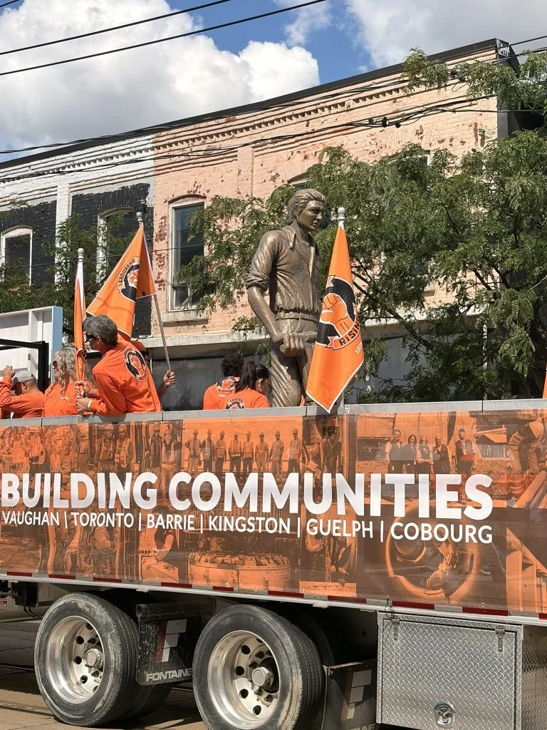 A flatbed truck carrying a Building Communities banner and orange union flags moves along a Toronto street past a statue.