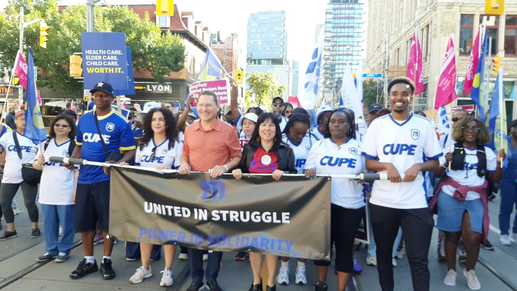 Marchers carrying a United in Struggle banner and union flags on a Toronto street, with downtown towers behind them.
