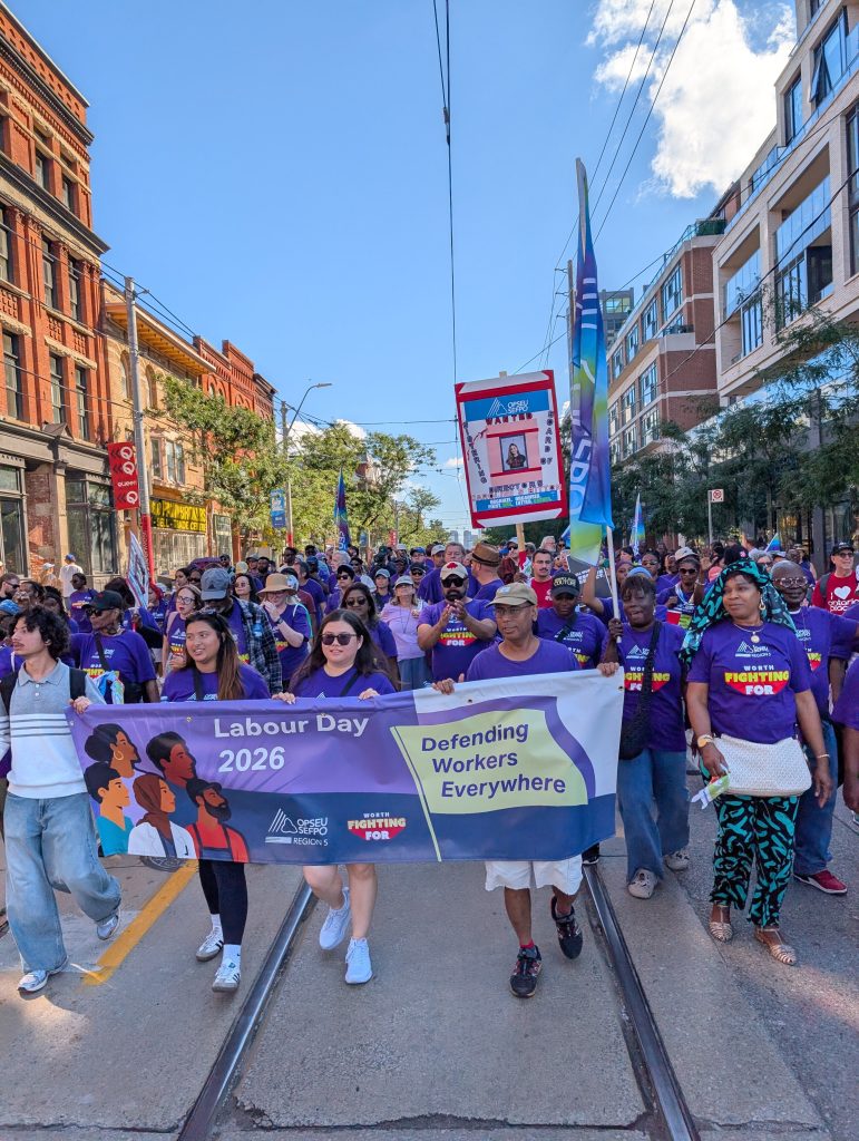 A wide column of marchers in purple fills a Toronto street with streetcar tracks, walking behind a banner reading Labour Day 2026, Defending Workers Everywhere.